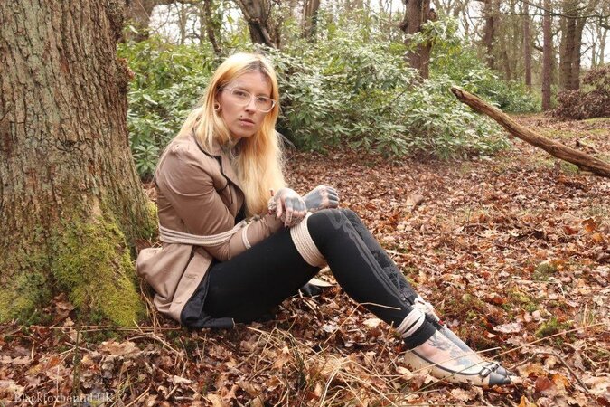 Girl with glasses can't stand up because she is hogtied by the tree