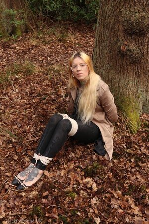Girl with glasses can't stand up because she is hogtied by the tree
