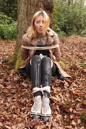 Girl with glasses can't stand up because she is hogtied by the tree